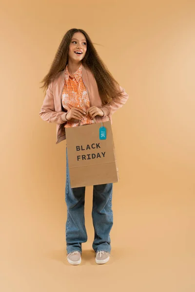 Amazed teenage girl holding paper bag with black friday lettering on beige — Stock Photo