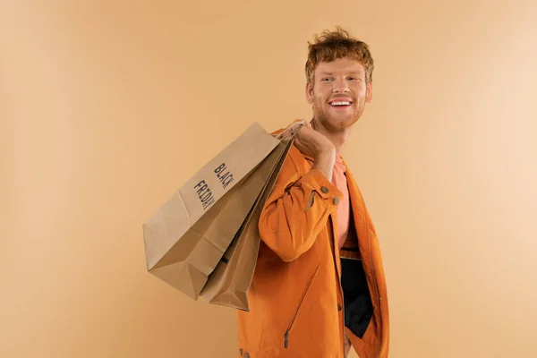 Cheerful young man with red hair holding shopping bags with black friday lettering isolated on beige — Stock Photo