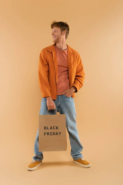 Positive young man with red hair holding shopping bag with black friday lettering and posing on beige — Photo de stock