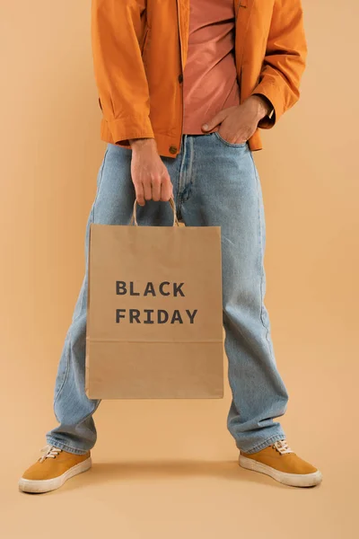 Cropped view of young man holding shopping bag with black friday lettering and posing on beige — Stock Photo