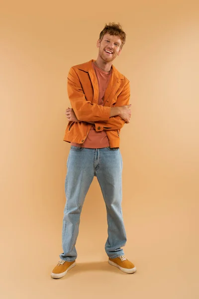 Full length of positive young man with red hair standing with crossed arms on beige — стоковое фото