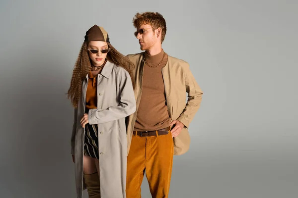 Young stylish couple in autumnal outfits and sunglasses posing on grey — стоковое фото