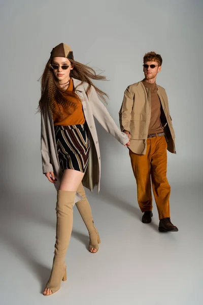 Full length of young couple in autumnal outfits and sunglasses holding hands while walking on grey — Photo de stock