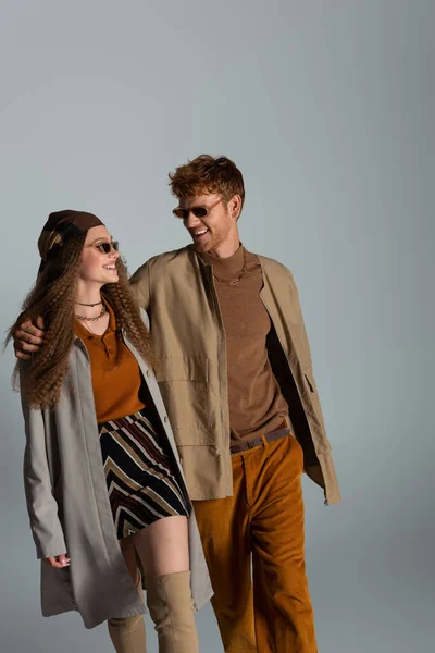 Cheerful young couple in autumnal outfits and sunglasses smiling isolated on grey — стоковое фото