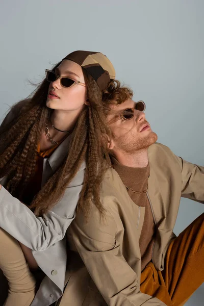 Young and stylish couple in sunglasses and autumnal outfits sitting together isolated on grey — стоковое фото