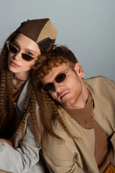 Молода і стильна пара в сонцезахисних окулярах і автентичних вбраннях дивиться на камеру ізольовано на сірому — стокове фото