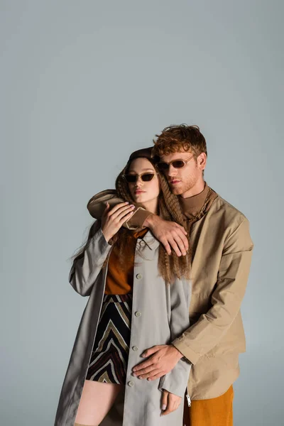 Young redhead man in sunglasses hugging girlfriend in headscarf isolated on grey — Foto stock