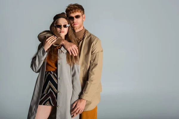Рудий чоловік в сонцезахисних окулярах обіймає стильну дівчину в спідниці і траншеї пальто ізольовано на сірому — стокове фото