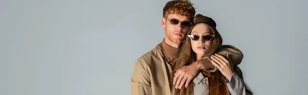 Redhead man in sunglasses hugging stylish girlfriend in trench coat isolated on grey, banner — Foto stock