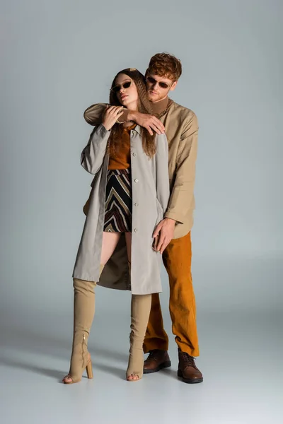 Full length of redhead man in sunglasses hugging stylish girlfriend in skirt and trench coat on grey — стоковое фото