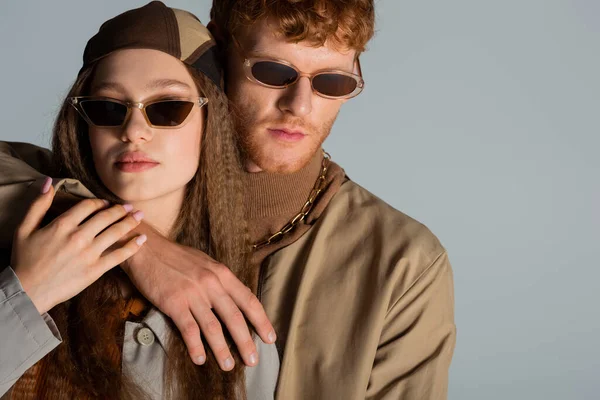Young redhead man in sunglasses hugging stylish girl in headscarf isolated on grey — стоковое фото