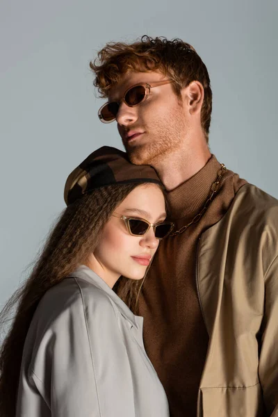 Young couple in sunglasses and autumnal clothes standing together isolated on grey - foto de stock