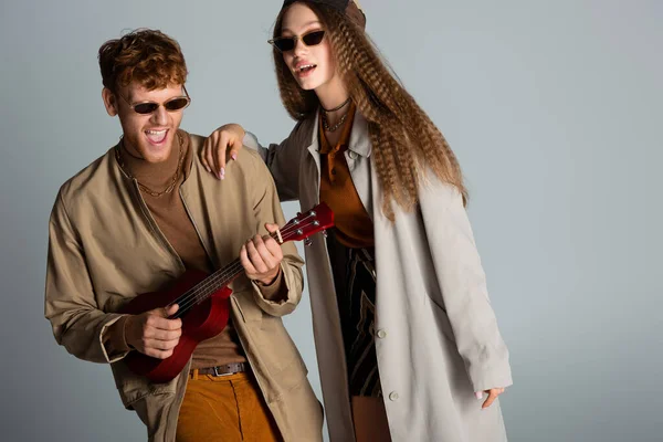 Amazed and redhead young man playing ukulele near girlfriend isolated on grey — Foto stock