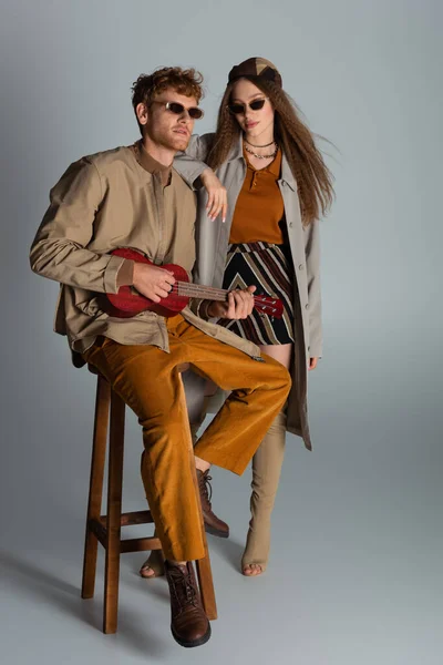 Full length of redhead young man playing ukulele and sitting on high chair near girlfriend on grey - foto de stock