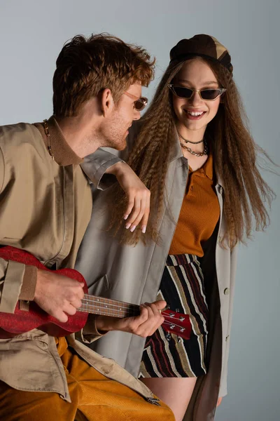 Redhead young man playing ukulele near smiling girlfriend isolated on grey — Stock Photo