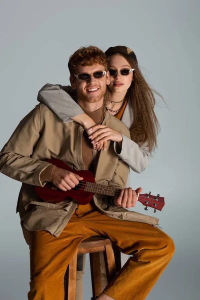 Happy young man playing ukulele while girlfriend in sunglasses hugging him isolated on grey — Stockfoto