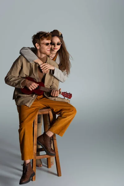 Щасливий молодий чоловік грає укулеле біля дівчини, сидячи на високому стільці на сірому — стокове фото