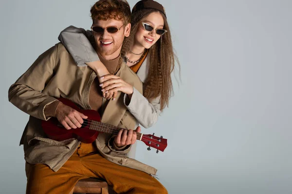 Redhead young man playing ukulele near happy girlfriend isolated on grey — Stockfoto