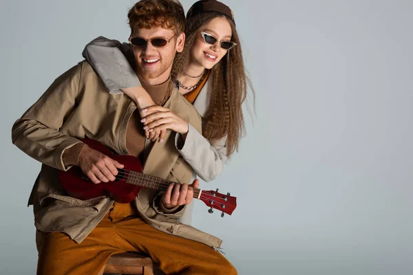 Redhead young man playing ukulele near happy girlfriend isolated on grey — стоковое фото