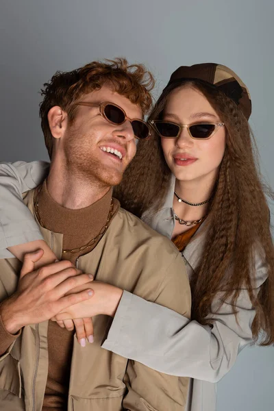 Stylish teenage girl in sunglasses hugging happy redhead boyfriend isolated on grey — стоковое фото