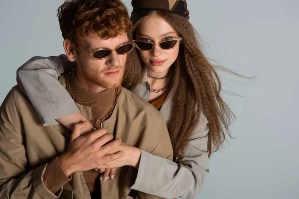 Teenage girl in sunglasses hugging stylish and redhead boyfriend isolated on grey — Stock Photo