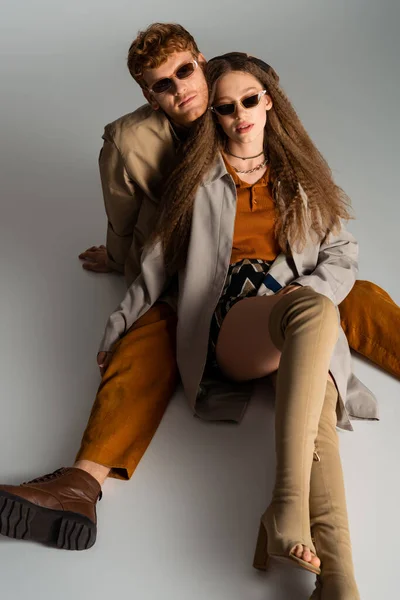 Stylish young couple in sunglasses and trendy outfits sitting on grey — Photo de stock