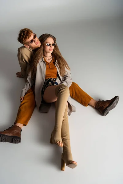 Young stylish couple in sunglasses and trendy outfits sitting on grey — стоковое фото