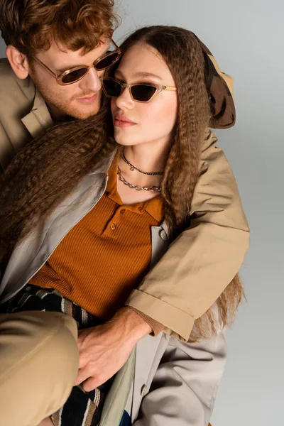 Stylish young man in trendy sunglasses hugging girlfriend isolated on grey — Stock Photo