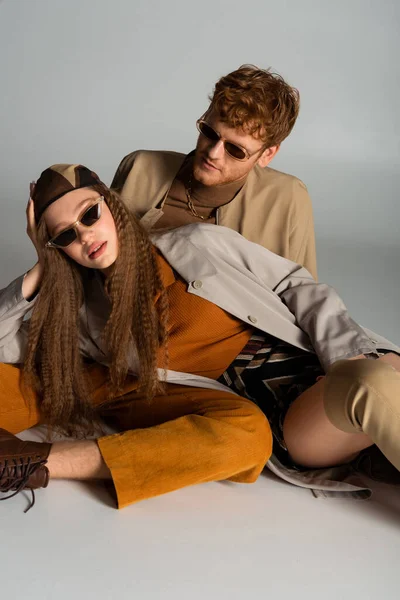 Stylish teenage model in sunglasses lying near redhead young man on grey — Stockfoto