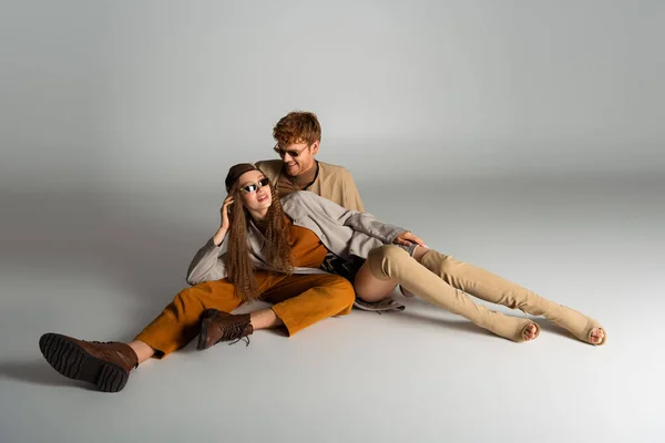 Stylish teenage model in sunglasses lying near happy young man on grey — Foto stock