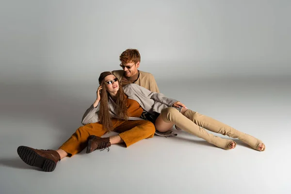 Stylish teenage model in sunglasses lying near happy young man on grey — Fotografia de Stock