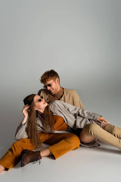 Stylish teenage model in sunglasses lying near cheerful young man on grey — Stock Photo