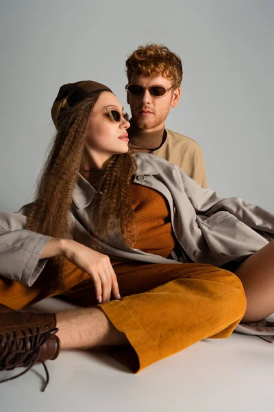 Redhead young man looking at stylish girlfriend in sunglasses posing on grey - foto de stock