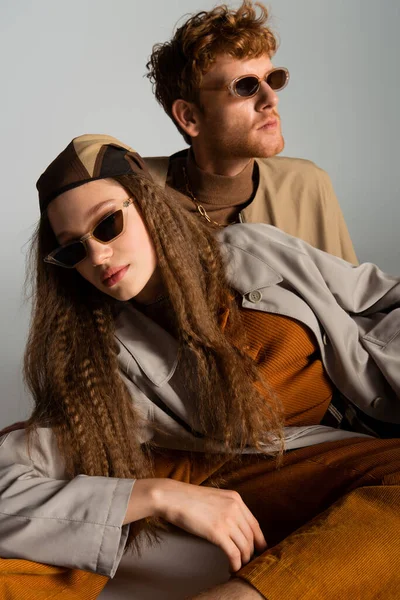 Redhead young man looking away near stylish model in sunglasses posing on grey - foto de stock