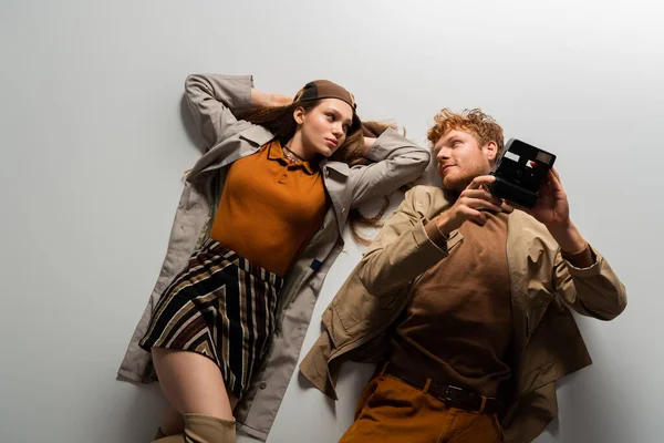 Top view of redhead young man in autumnal outfit holding vintage camera and lying together with girlfriend on grey — Photo de stock