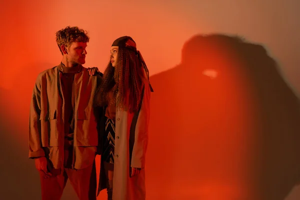 Young and stylish couple in autumnal outfits looking at each other on red background — Stock Photo