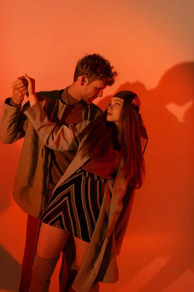 Young stylish models in autumnal outfits looking at each other and holding hands on red — Photo de stock