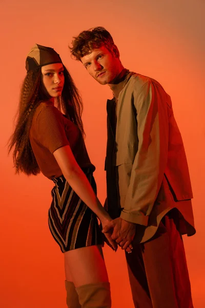 Stylish couple in autumnal outfits holding hands and looking at camera on red — Fotografia de Stock