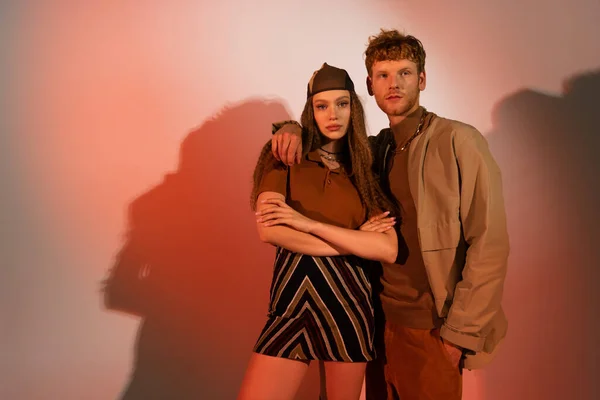 Young redhead man standing with hand in pocket near stylish girl in headscarf and skirt on red background — Foto stock