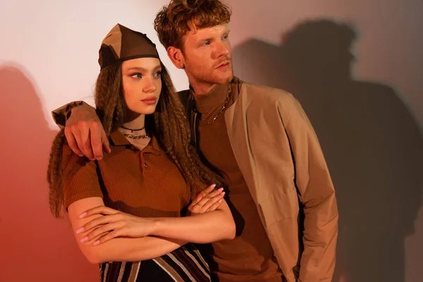 Young redhead man hugging stylish model in headscarf standing with crossed arms on red — Stockfoto