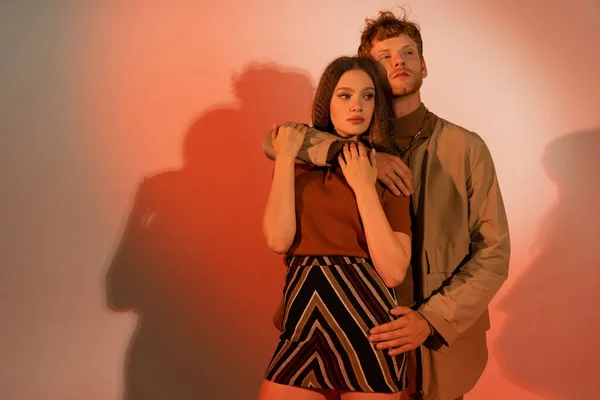 Redhead young man hugging stylish girlfriend in skirt on red background — стоковое фото