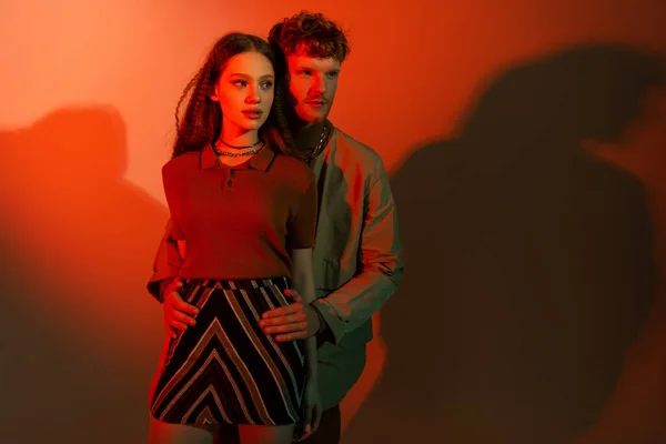 Young redhead man standing and hugging hips of stylish girl in skirt on red background — Photo de stock