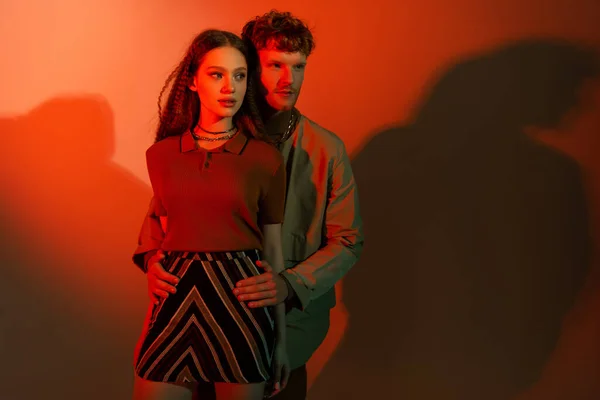 Young redhead man standing and hugging hips of stylish girl in skirt on red background — Foto stock