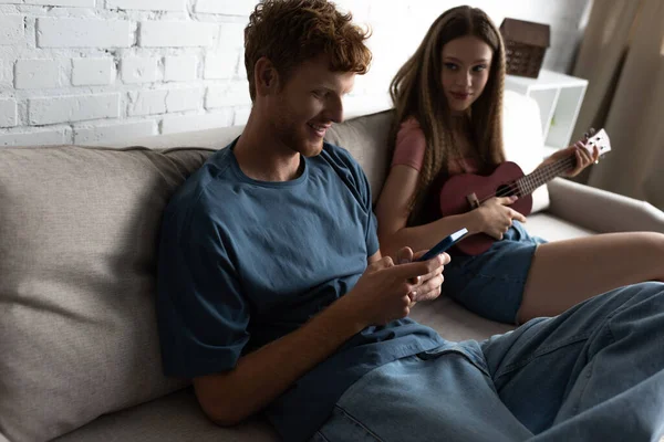 Teenage girl playing ukulele and looking at happy boyfriend using smartphone while sitting on couch — стоковое фото