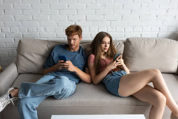 Young couple using smartphones while sitting on couch in living room — Stockfoto