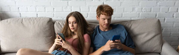 Young couple using smartphones while sitting on couch in living room, banner — Photo de stock