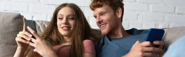 Young couple using mobile phones while sitting on couch in living room, banner - foto de stock