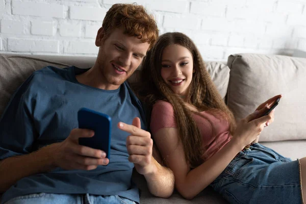 Young man pointing at mobile phone while sitting on couch with happy girlfriend in living room — Fotografia de Stock