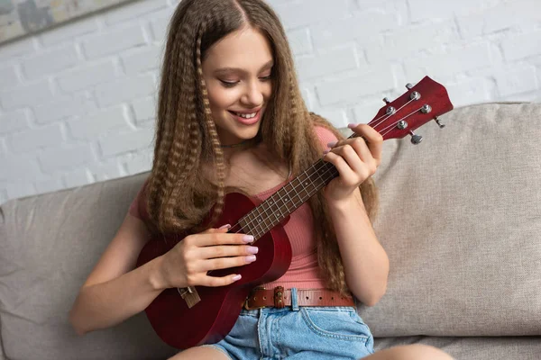 Joyful teenage girl in casual clothes playing ukulele while sitting on couch in living room — Photo de stock