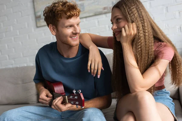 Curly and happy young man playing ukulele near girlfriend in living room — Fotografia de Stock